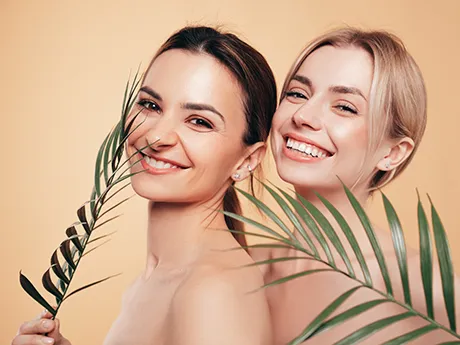 Natürliche Hautpflege mit PMA-Zeolith – CARE Naturkosmetik für Haut und Zähne Zwei lächelnde Frauen mit gesunder Haut stehen vor beigem Hintergrund und halten grüne Palmblätter. Das Bild symbolisiert natürliche Schönheit, Pflege und Hautgesundheit mit PMA-Zeolith.